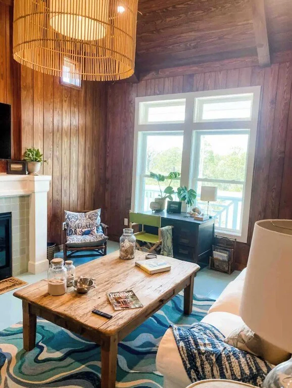 A cozy living room with wooden walls, large windows, and a rustic wooden coffee table. There is a palm plant on a black desk by the window, a decorative chair with a patterned cushion, a white fireplace with a plant on top, a beige sofa with a patter