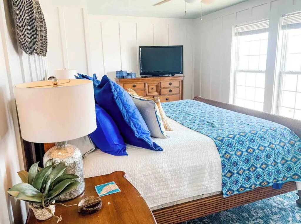A bedroom with a large bed covered in a white quilt and a blue patterned bed runner. The headboard has blue pillows, and there are beige pillows with colorful accents. Next to the bed, a wooden nightstand holds a glass table lamp with a white shade,