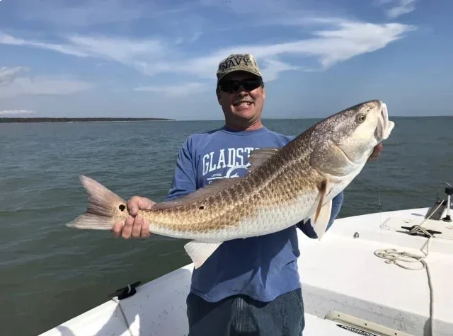 Man in sunglasses holding a large striped fish on a boat, with water and sky in the background.