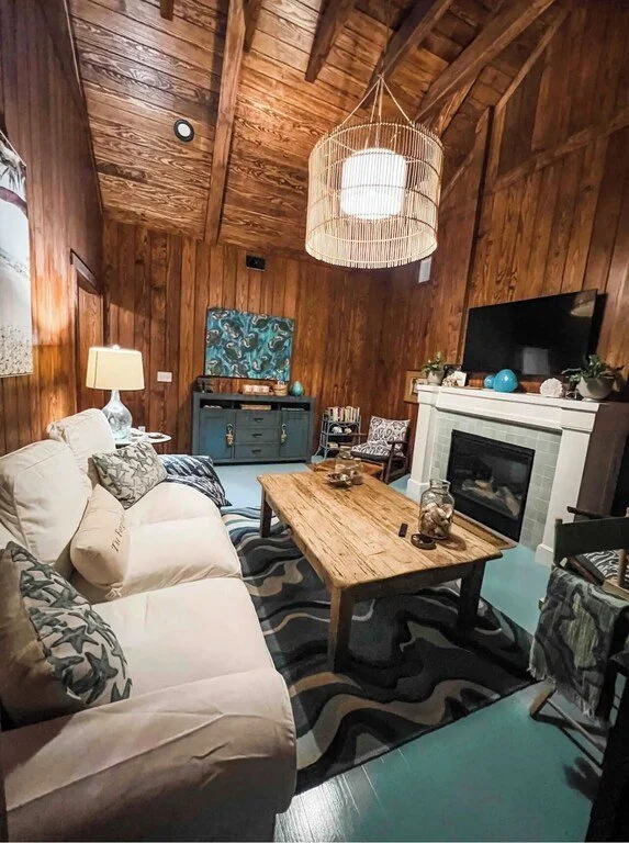 Cozy living room with wood-paneled walls and ceiling, a white sofa with decorative pillows, a wooden coffee table, a flat-screen TV above a white fireplace mantel, a chandelier, a side table with a lamp, and various decorative items, creating a warm