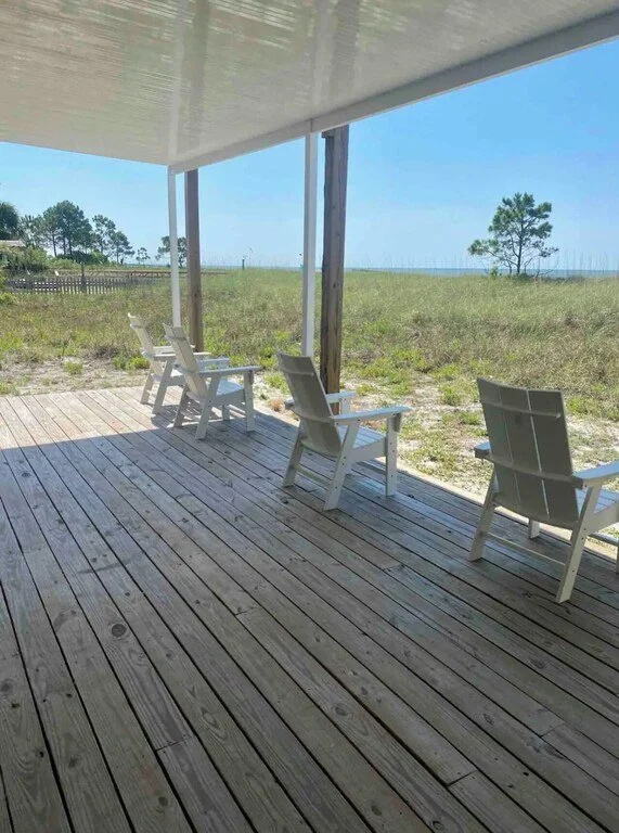 Three white Adirondack chairs on a wooden porch overlooking grassy dunes and a few trees.