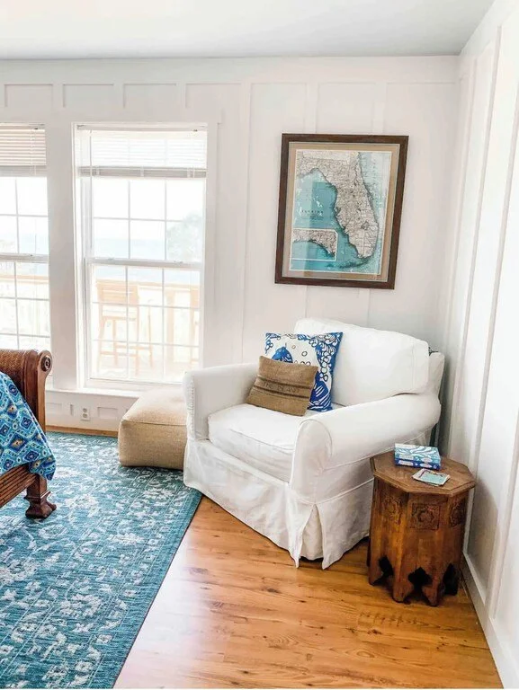 Living room corner with white armchair, wooden side table, framed map on the wall, and large windows with a balcony outside.
