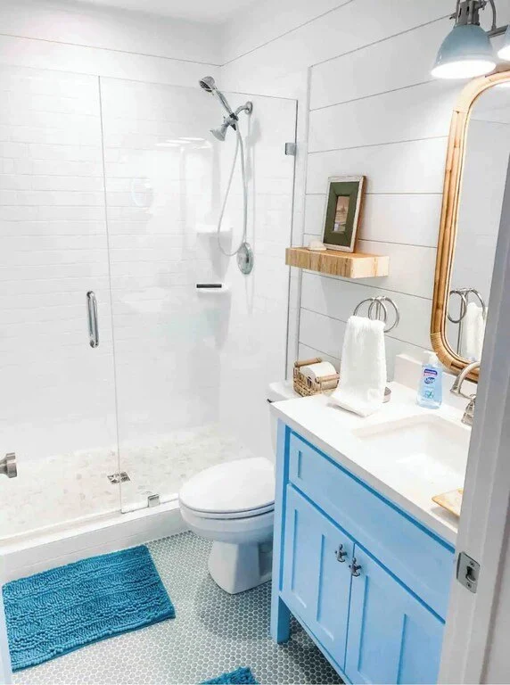 A small bathroom with a glass shower enclosure, a white toilet, and a blue vanity with a mirror. There is a wooden shelf with a framed picture above the toilet, and a towel hanging on a ring. A blue bath mat is on the tile floor.