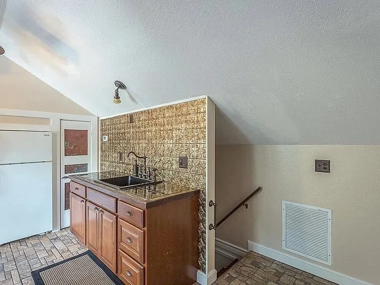 Small kitchen area with a brown wooden cabinet, tiled backsplash, black sink, and a light-colored textured wall. A stairway with a wooden handrail leads down from the kitchen.