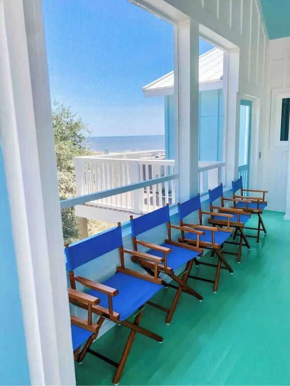 Beachside porch with six blue director's chairs facing large windows, overlooking a sandy beach and ocean under a clear blue sky.