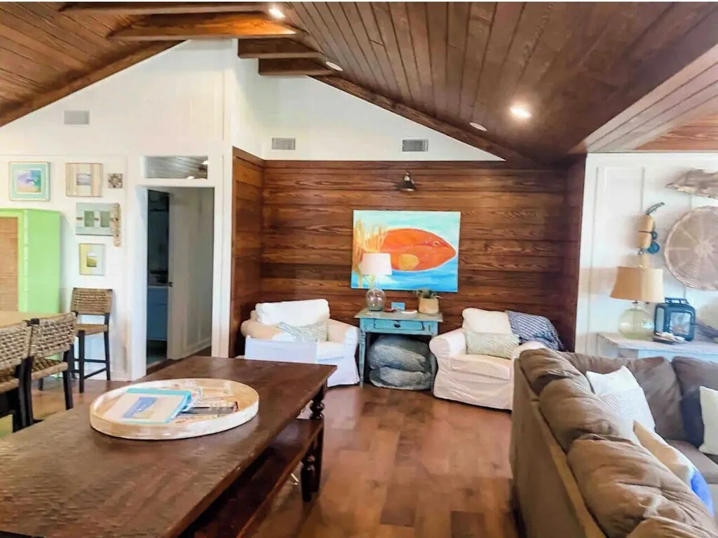 Living room with wooden ceiling and walls, featuring a colorful fish painting, two white armchairs, a dark blue console table, a brown sofa, and various decorative items.