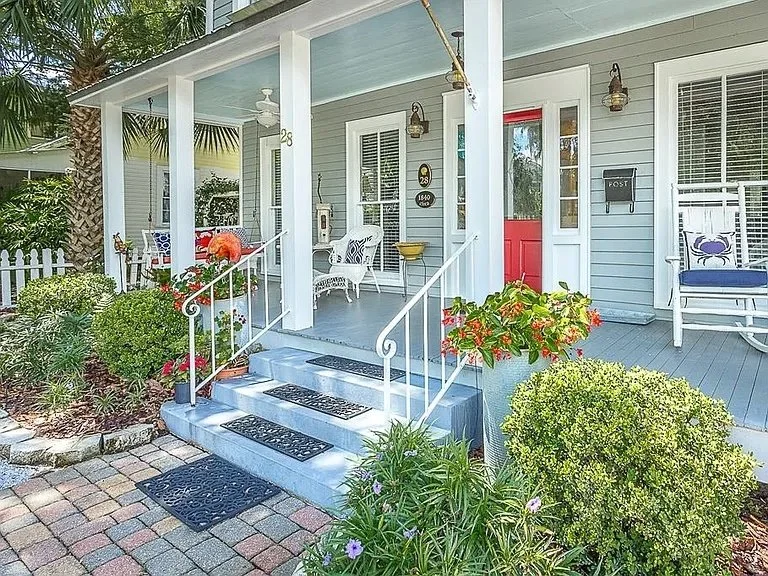 Front porch of a house with a small garden, potted plants, and patio furniture, including a bench and chairs, with white railings and steps leading to the door, decorated with outdoor lighting.