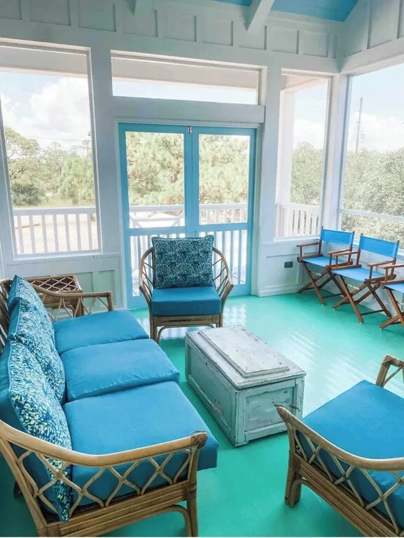 Bright porch with white walls and mint green flooring, furnished with a blue cushioned rattan sofa, matching armchair, three blue folding chairs, and a white distressed coffee table.