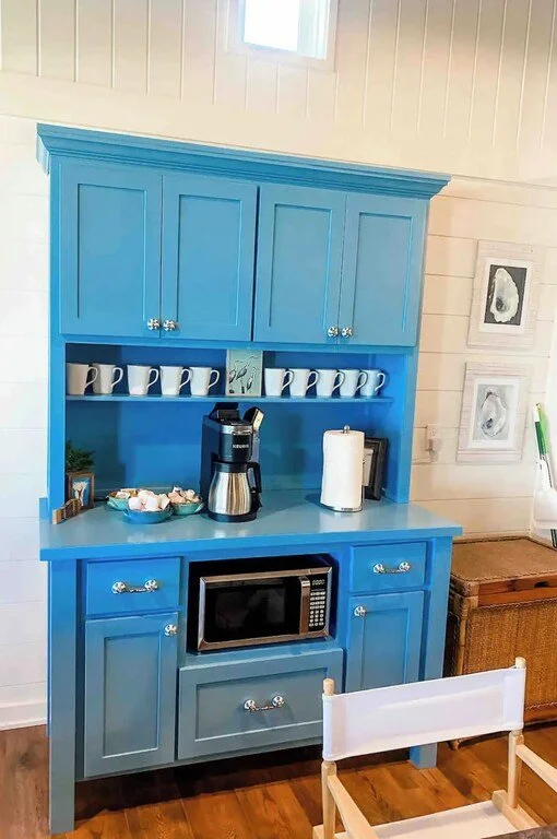 Bright blue kitchen cabinet with upper and lower cabinets, open shelf with coffee mugs, coffee maker, paper towel holder, microwave, and a wooden chest nearby.