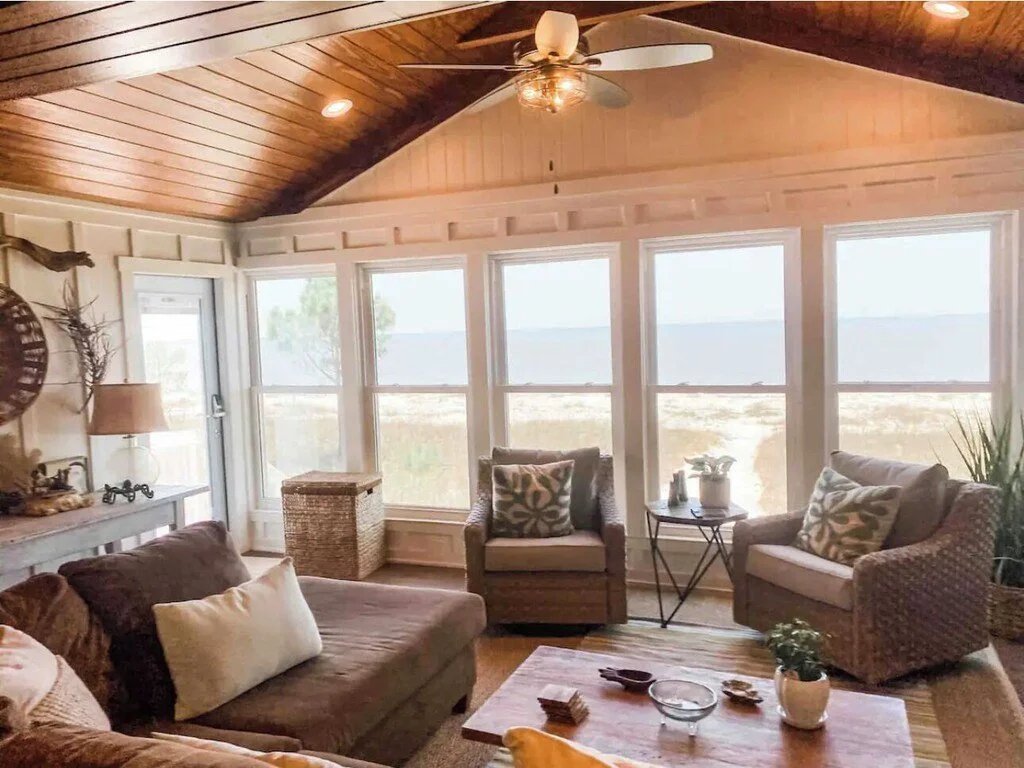 Living room with large windows overlooking a beach, featuring a brown sofa with white pillows, two wicker armchairs with patterned pillows, a wooden coffee table, a table with potted plants, and a ceiling fan with a light.