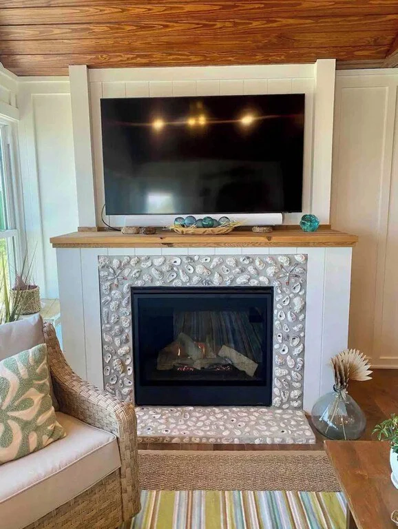 Living room with a flat-screen TV mounted above a fireplace with a shell design frame, and a wooden mantel with decorative objects. Part of a wicker chair with a patterned pillow, a glass vase with dried flowers, and a colorful striped rug are also v