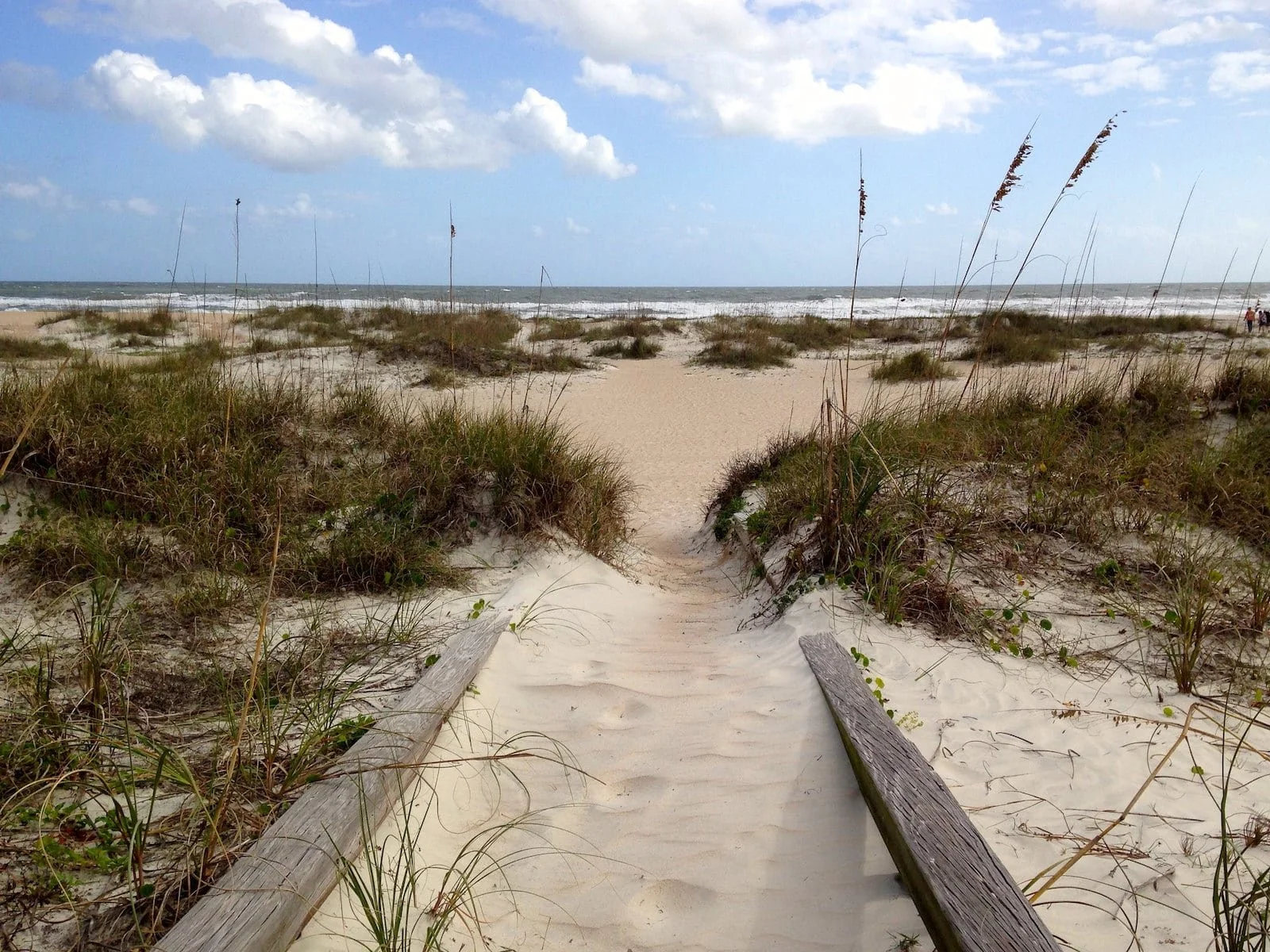 Wooden pathway leading through sandy dunes with beach grass towards the ocean, cloudy sky overhead.