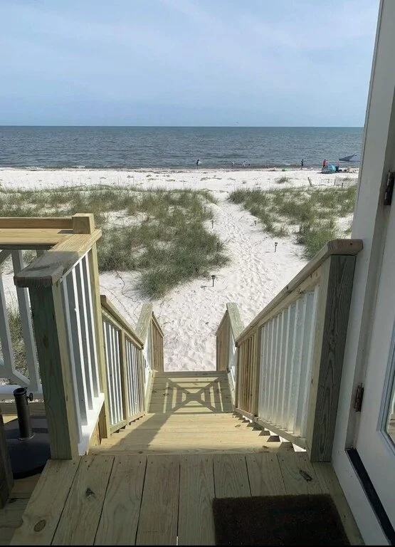 View from a beach house doorway looking down a wooden staircase to sandy beach with patches of grass, ocean waves, and a clear sky.