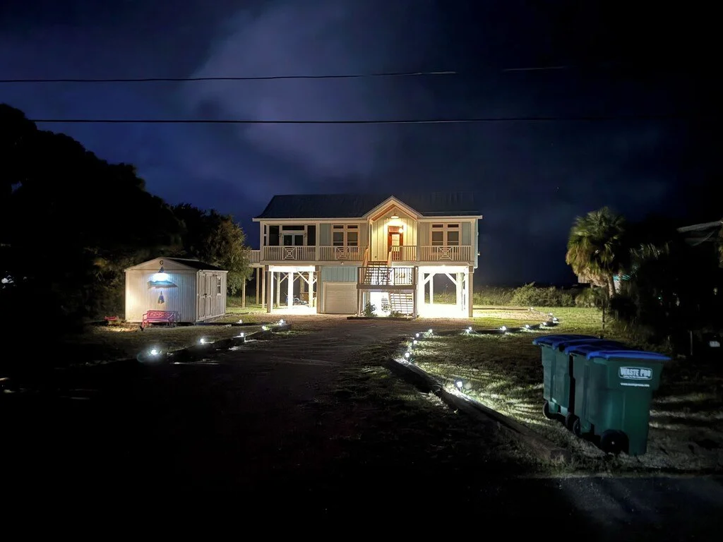 A two-story house elevated on stilts at night, illuminated by outdoor lights along a pathway, with a small white shed nearby and a green trash bin in the front yard, under a dark sky with clouds.