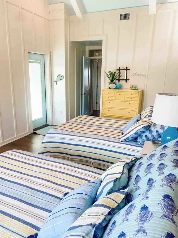 A bedroom with two beds with striped bedding, a yellow dresser with a plant and decorative objects on top, a white lamp, and light-colored walls with wood paneling. There's a glass door on the left and a door leading to another room or closet.
