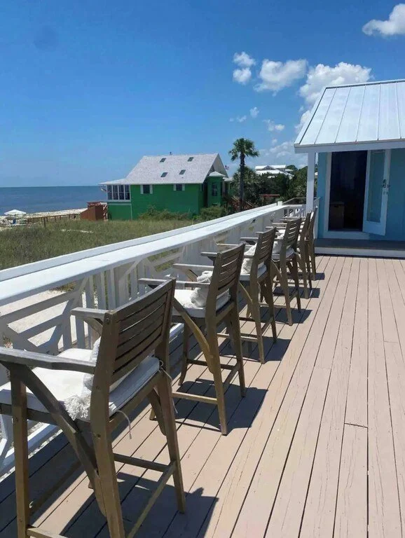 Balcony with seven wooden chairs overlooking the beach, ocean, and a green house with a white roof.