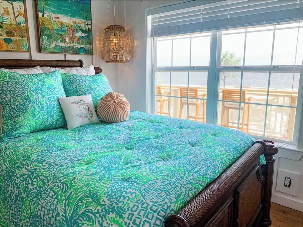 A bedroom with a large bed covered in green and blue patterned bedding, decorative pillows, and ocean view through large windows with a balcony and outdoor seating.