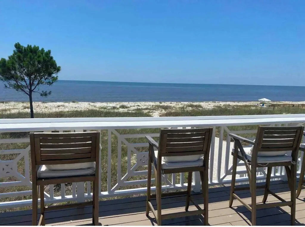 View from a porch with three wooden chairs overlooking a beach with sand, a lone tree, and the ocean under a blue sky.
