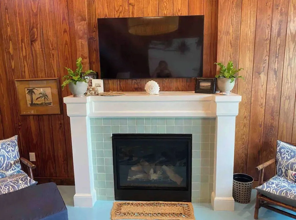 Living room fireplace with a flat-screen TV mounted above and wooden panel walls. Decor includes plants, framed pictures, and a white seashell on the mantel. Two chairs with patterned cushions are on either side.