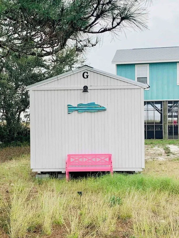 White shed with a blue fish decoration on the front and a pink bench in front. A black letter G at the top of the shed and a small black light below the roof. Green grass and a tree with branches over the shed. A turquoise building in the background.