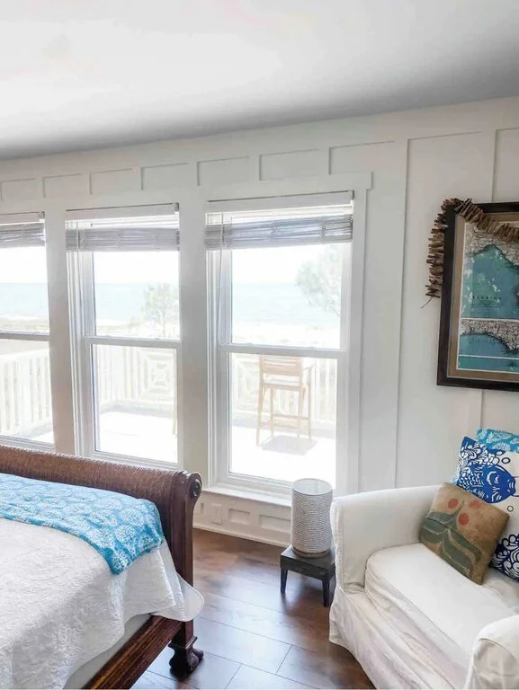 A bright bedroom with three large windows overlooking a balcony with outdoor chairs. The room has white walls, dark wood flooring, a white sofa with pillows, a small side table, and wall decor.