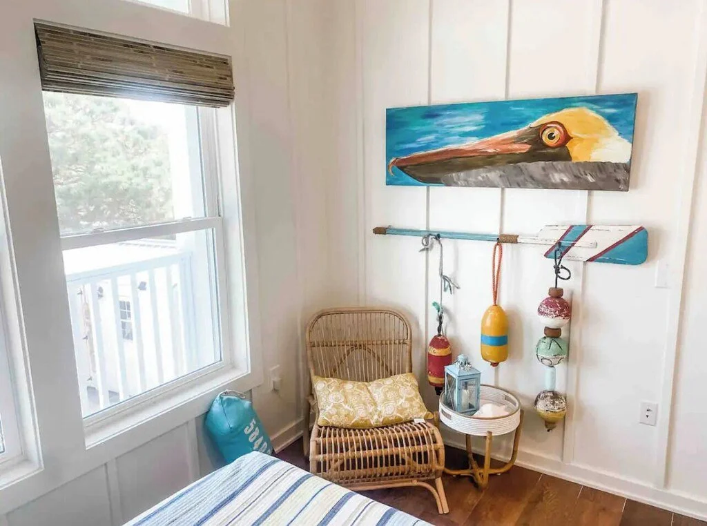 A cozy corner with a rattan chair and a yellow patterned cushion next to a small white round side table holding a lantern. Decor includes a large painting of a pelican head above a wooden rod with hanging buoy and lanterns, next to a window with a vi