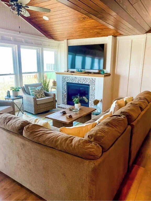Living room with beige couch, armchair, wooden coffee table, fireplace with decorative tile, wall-mounted TV, large windows with view, and wooden ceiling.