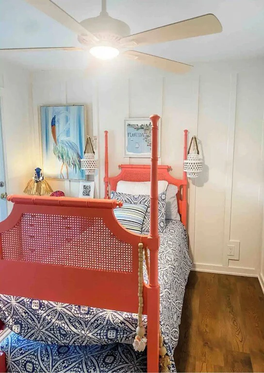 A bedroom with a coral-colored four-poster bed, striped and patterned bedding, and tropical artwork on the wall. There is a ceiling fan with a light, and two woven hanging baskets on the headboard. The wall is paneled, and the floor is wood.