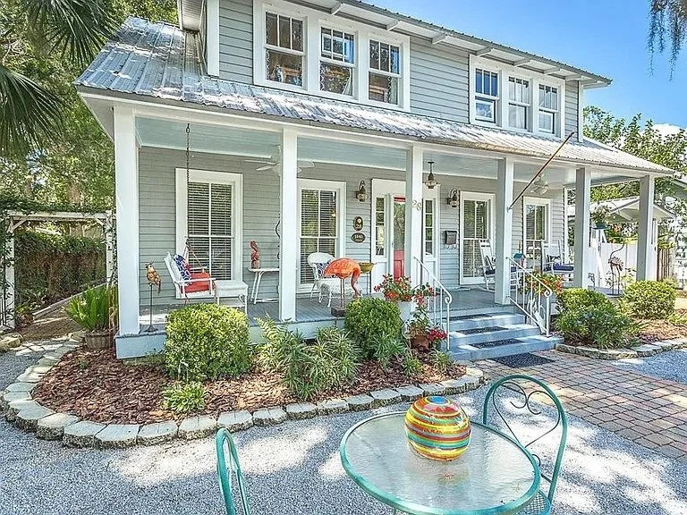 Front porch of a two-story house with white siding, blue trim, and rocking chairs. The house has hanging swings on the porch and a brick pathway leading to the steps. There is a garden with bushes and plants in front and a colorful striped vase on a