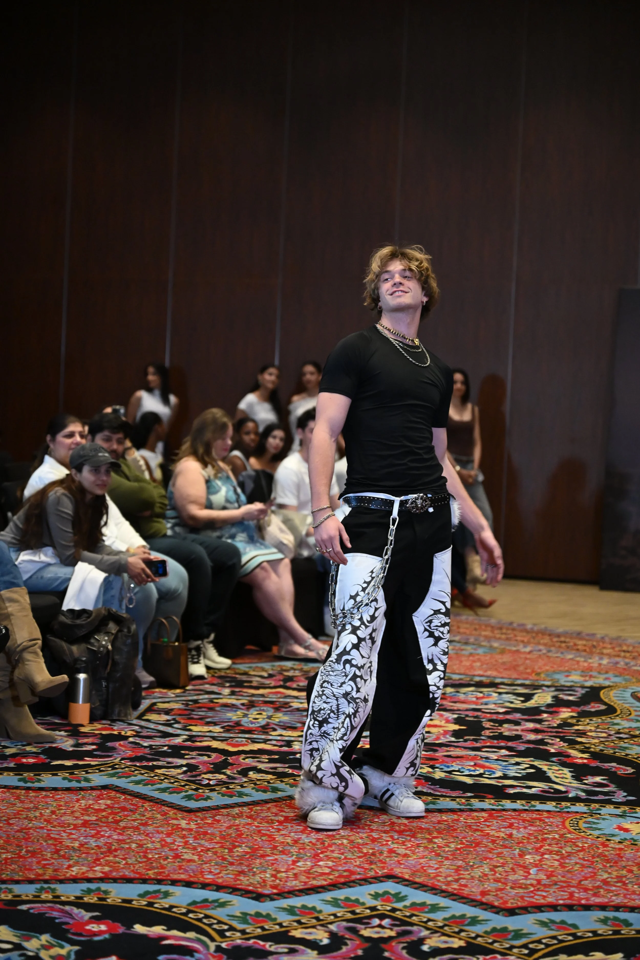 A male model walking on a runway during a fashion show, with an audience seated on both sides, some taking photos.
