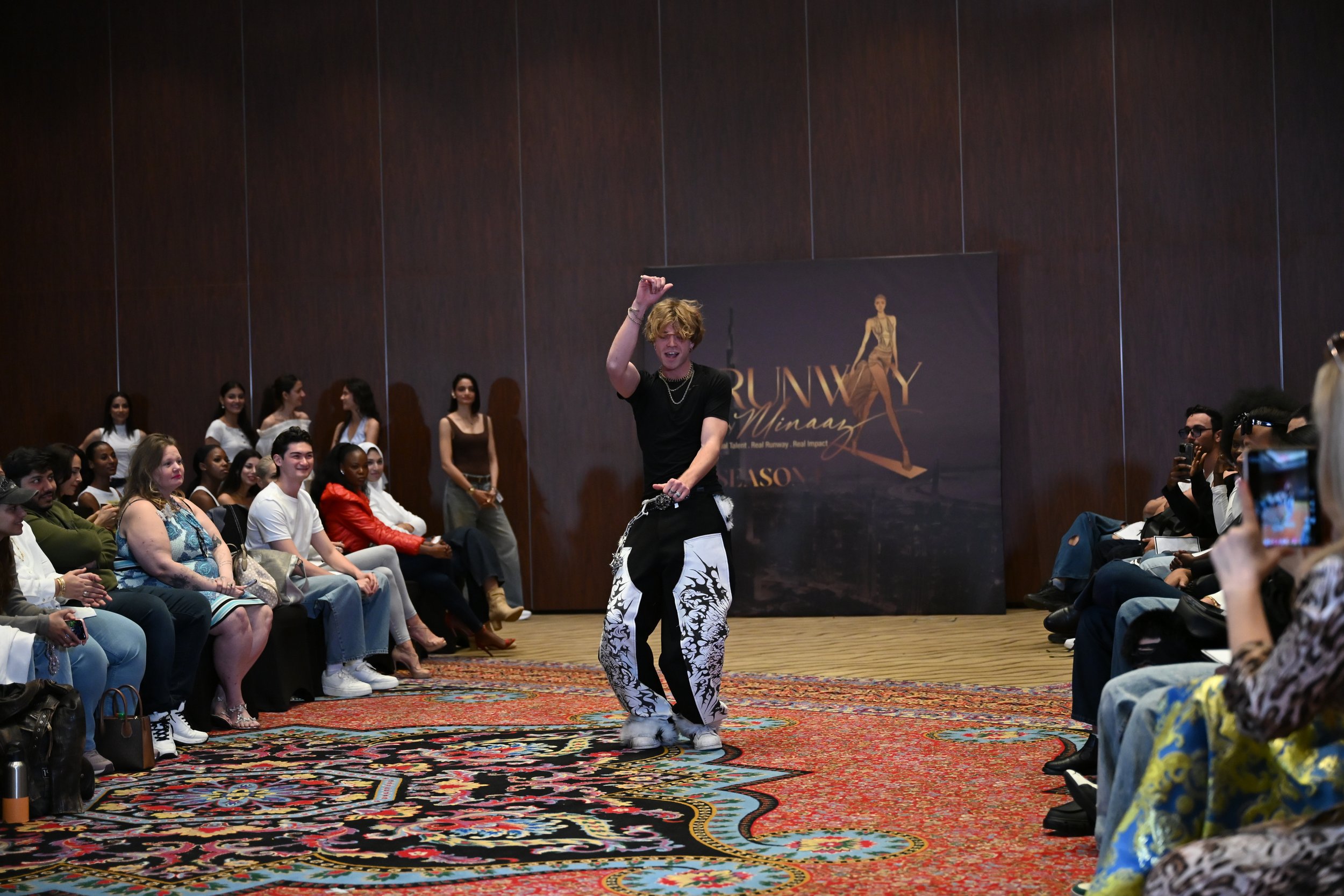 A young man with blond hair dancing on a runway at a fashion show, surrounded by seated audience members taking photos, with a dark wooden wall and a promotional poster in the background.