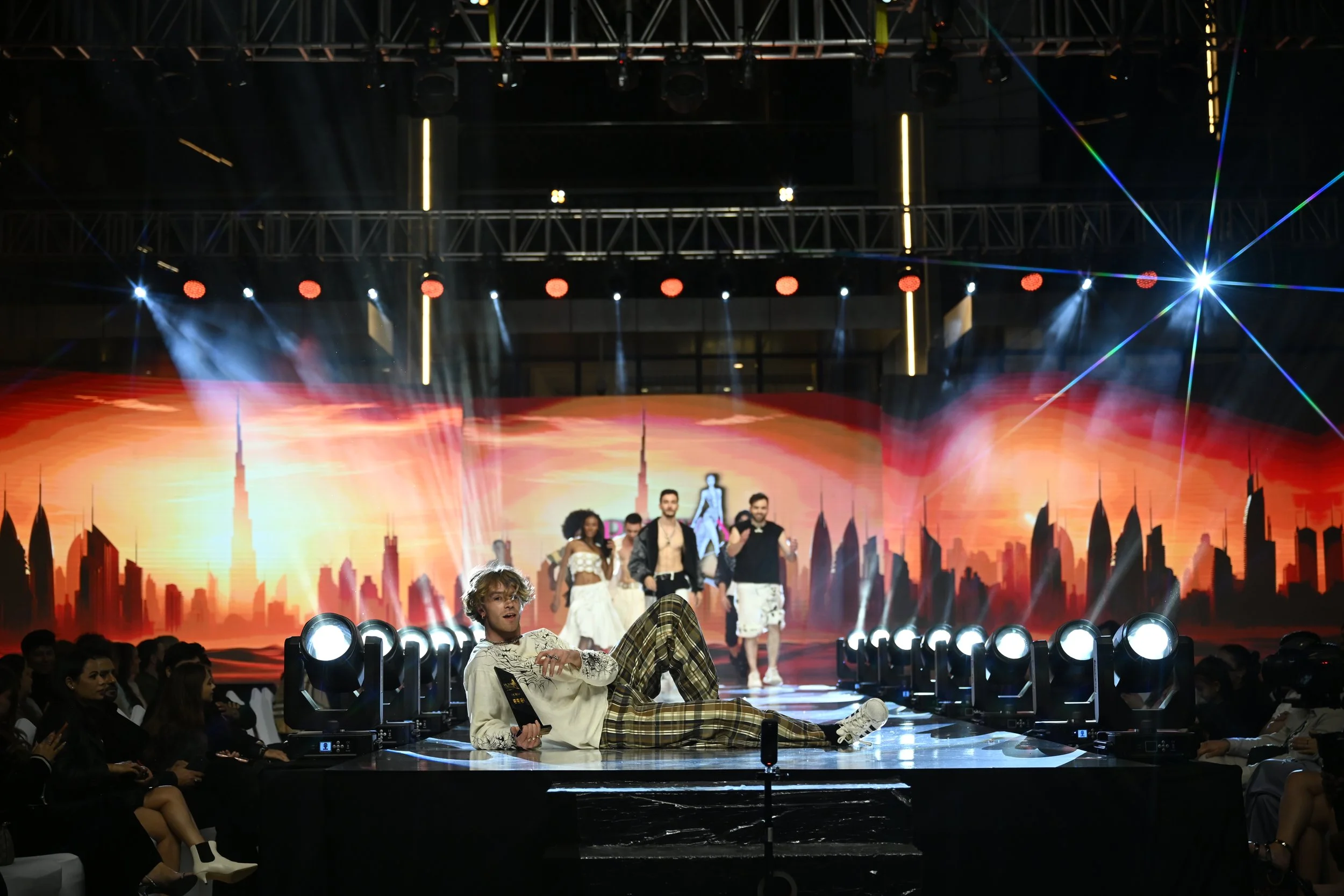 A fashion show runway with models walking and one model mid-stride, while a person lounges on the stage posing with one leg extended and holding a remote or award, surrounded by bright stage lights and a digital cityscape background.