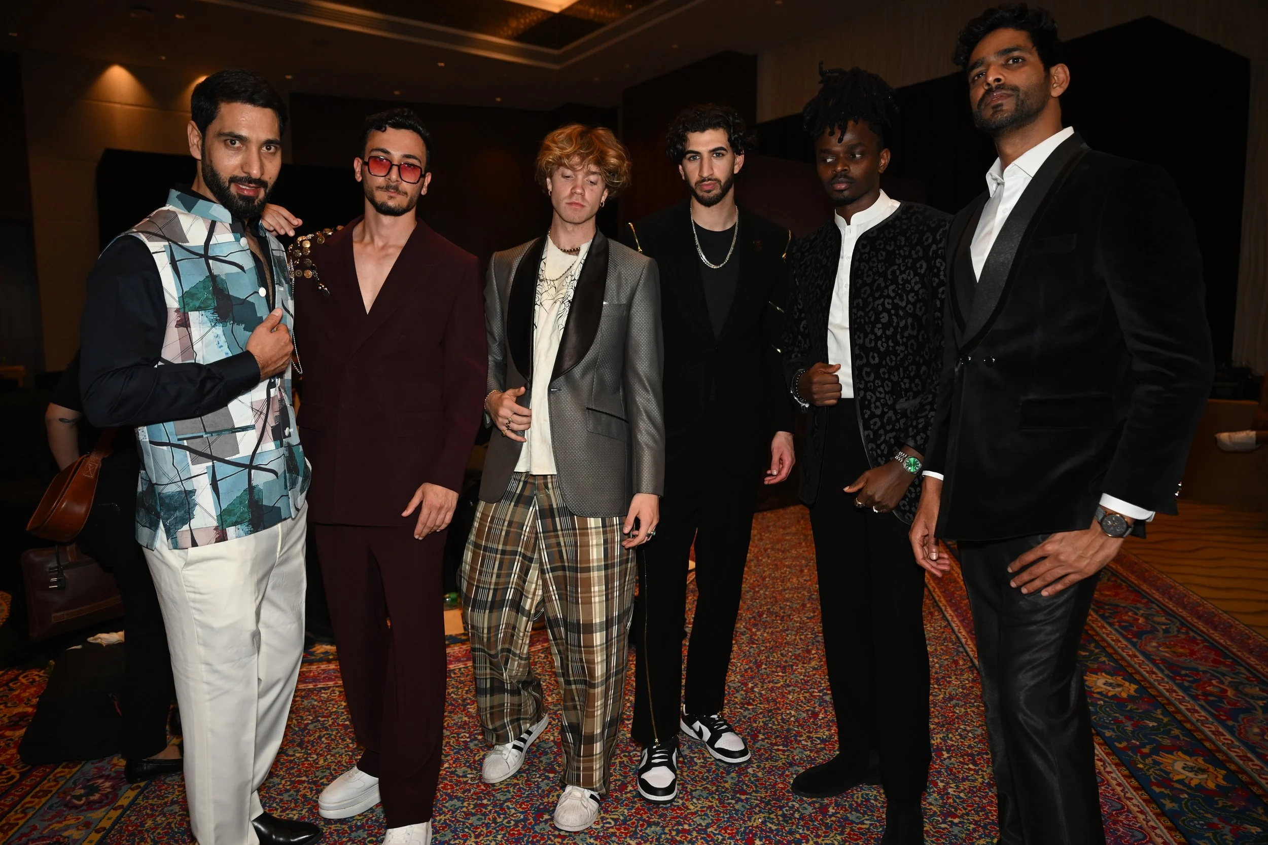 A group of six men posing together indoors, dressed in stylish, fashionable clothing.