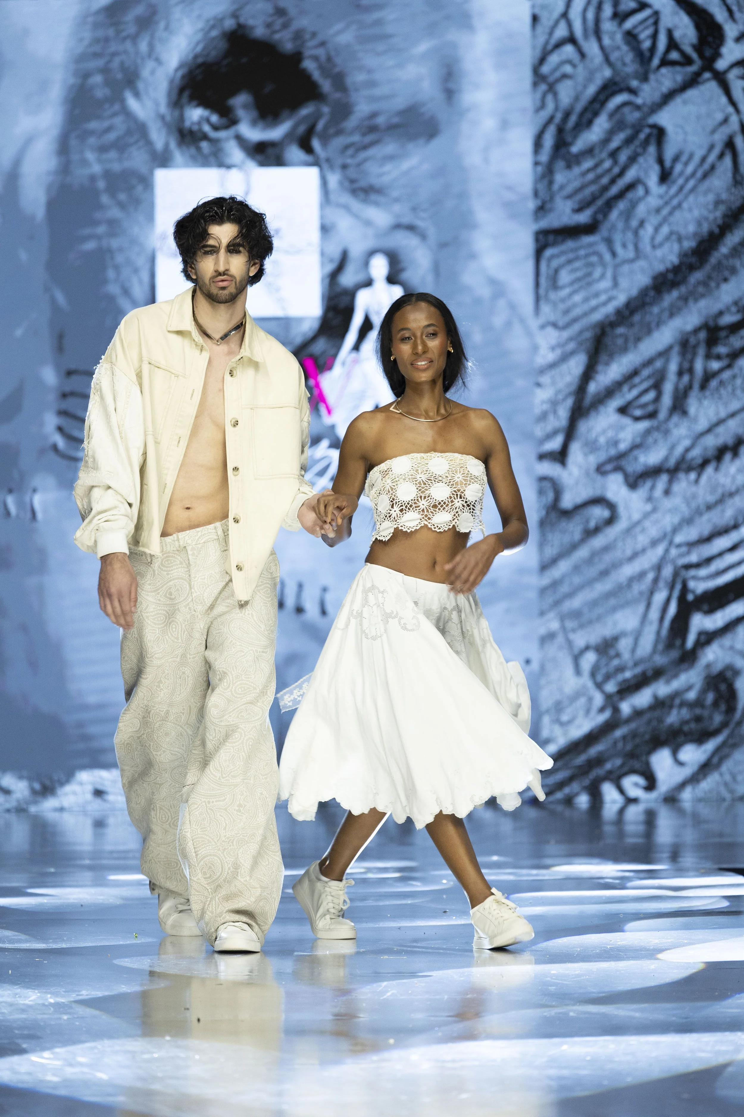 Models walking down runway during a fashion show, with a large digital screen in the background displaying artistic imagery.