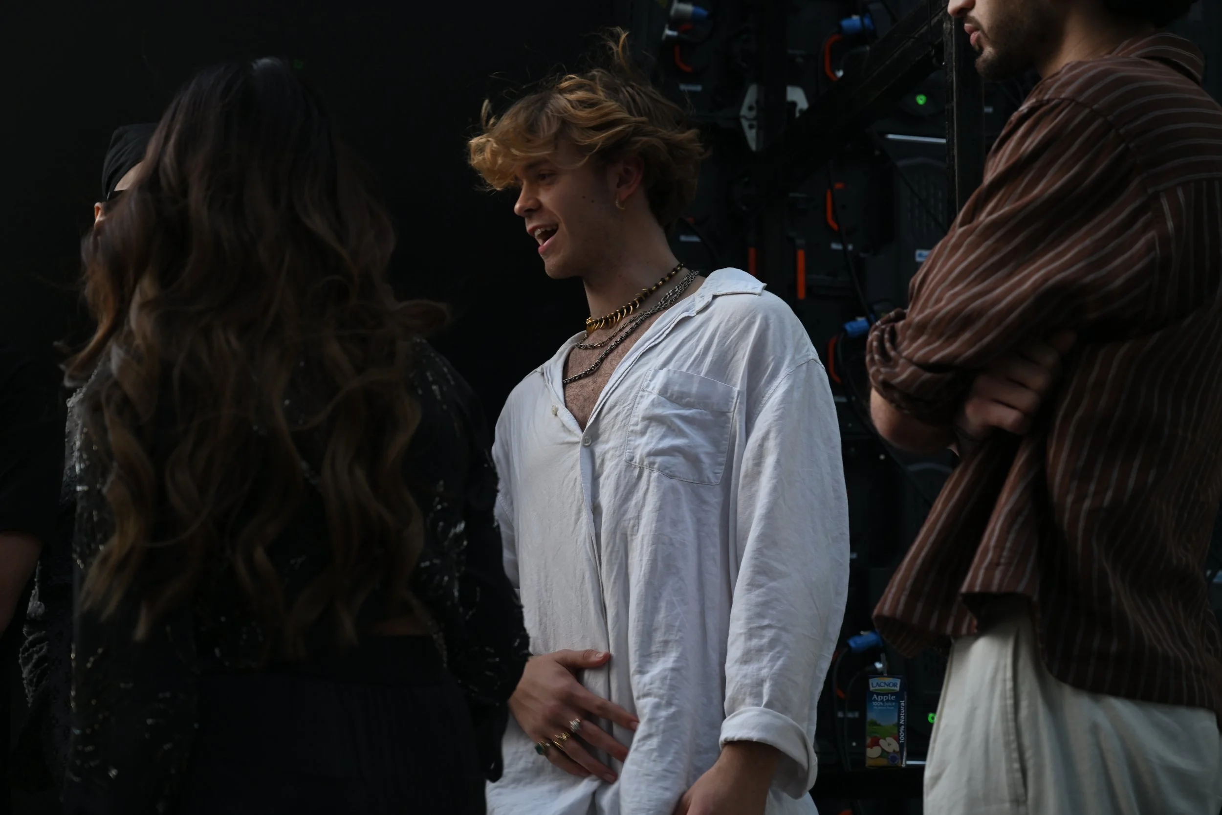 Three young people in conversation, two women and a man, in a dimly lit room with technical equipment in the background. The woman on the left has long wavy hair, the man in the middle has curly blond hair and is wearing a white shirt and layered nec