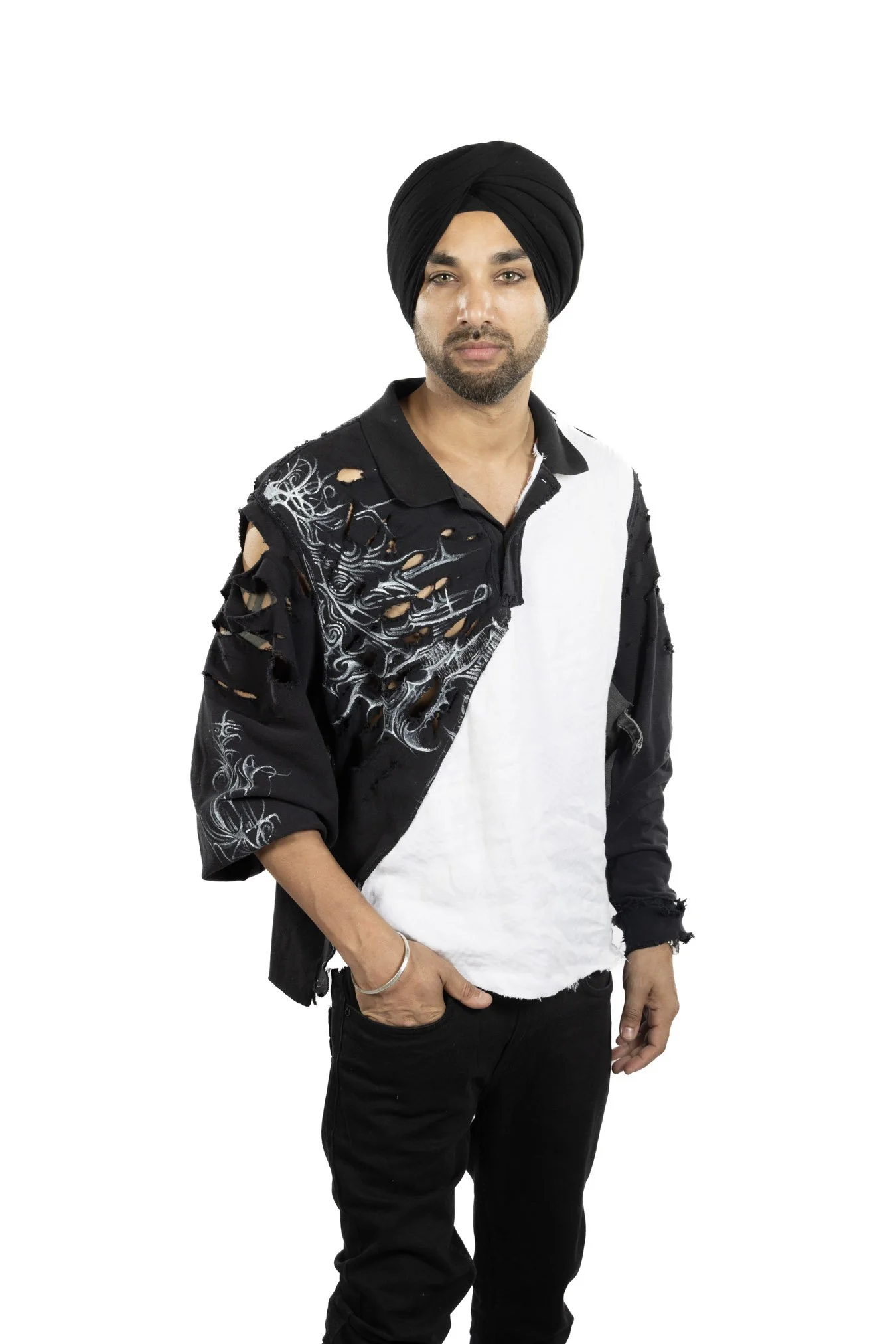 A man wearing a black turban, black ripped jacket with white design, and black pants poses with one hand in his pocket against a white background.