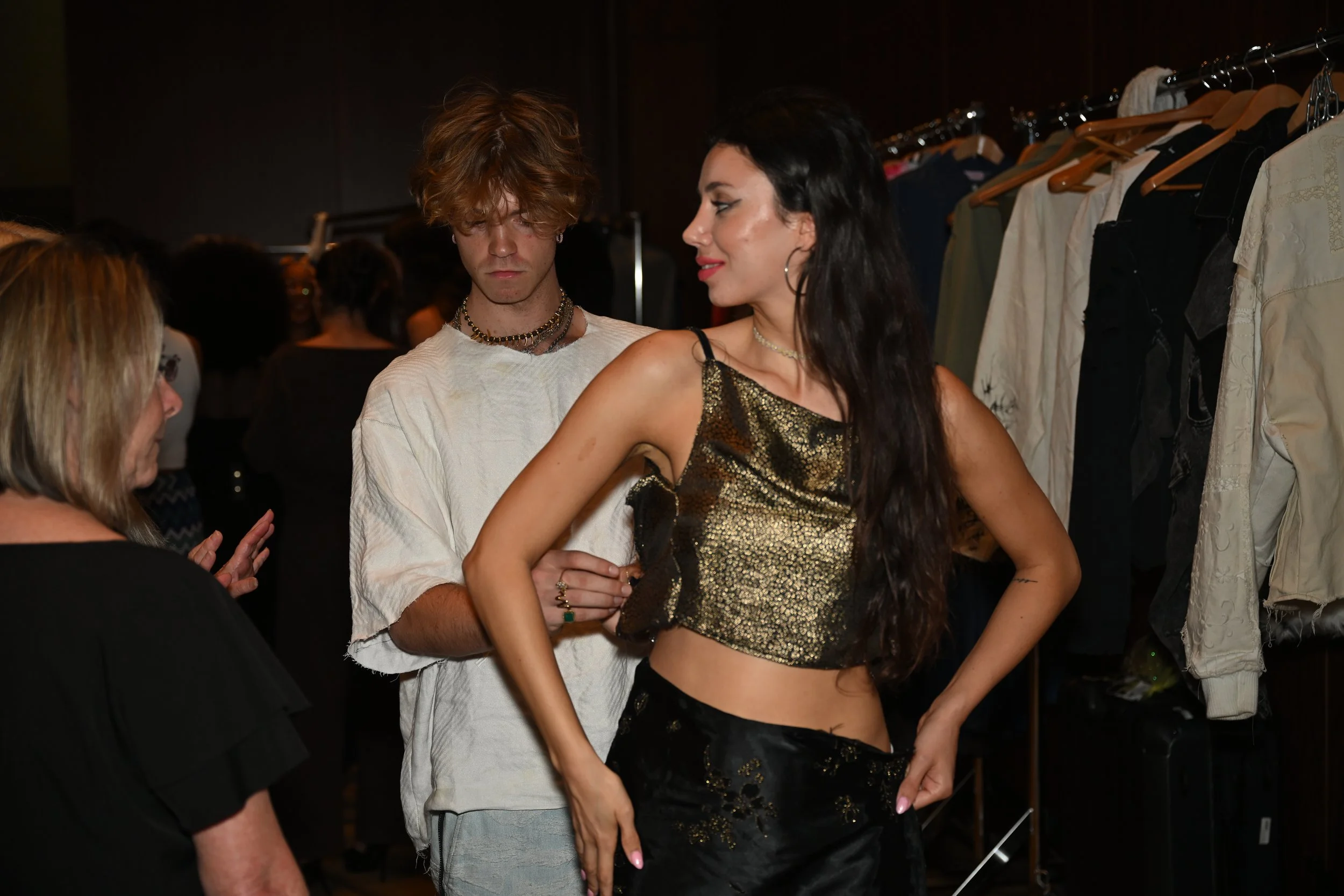 Model in black and gold outfit trying on clothes at a fitting while two women assist her in a backstage area