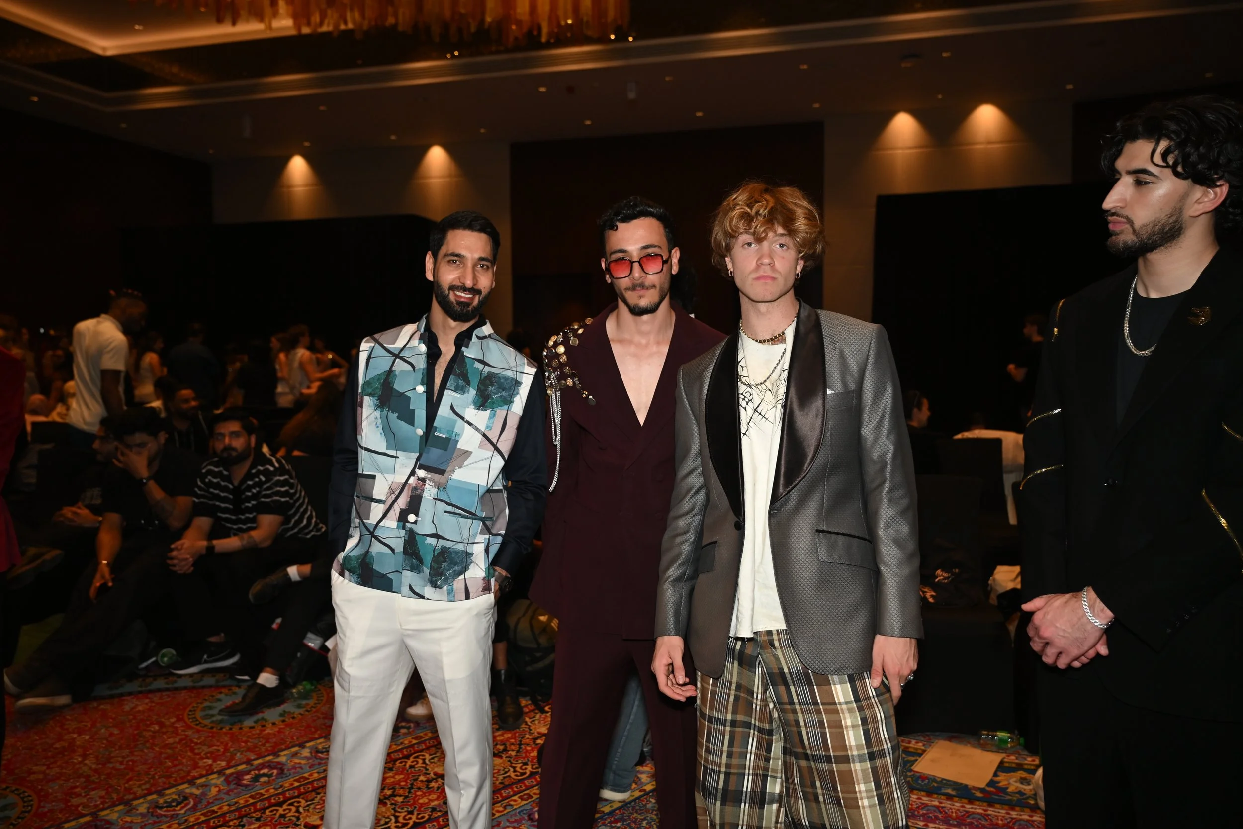 Four men standing together in stylish clothing at an indoor event with many people seated and standing in the background.