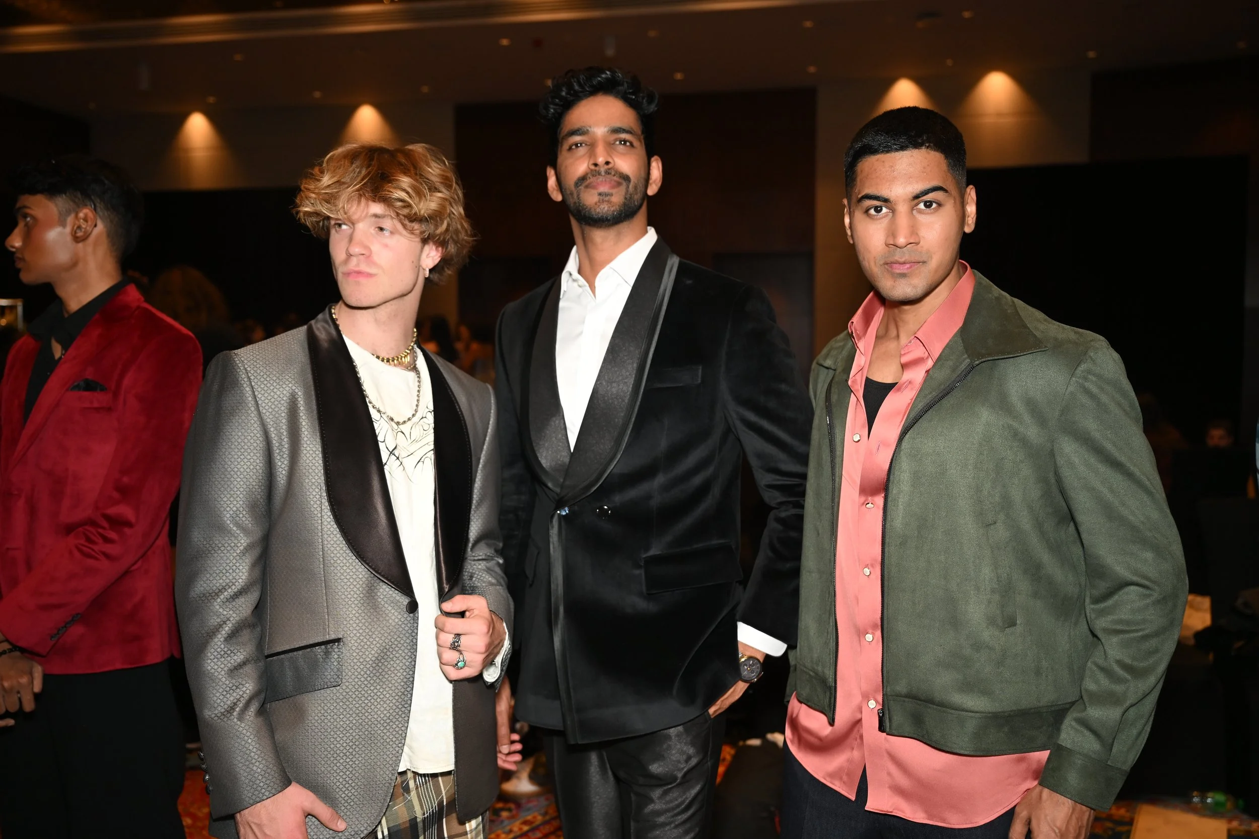 Three young men in fashionable clothing standing together at an indoor event, with one in a gray blazer, another in a black velvet tuxedo, and the third in a green jacket and pink shirt. People and warm lighting are in the background.