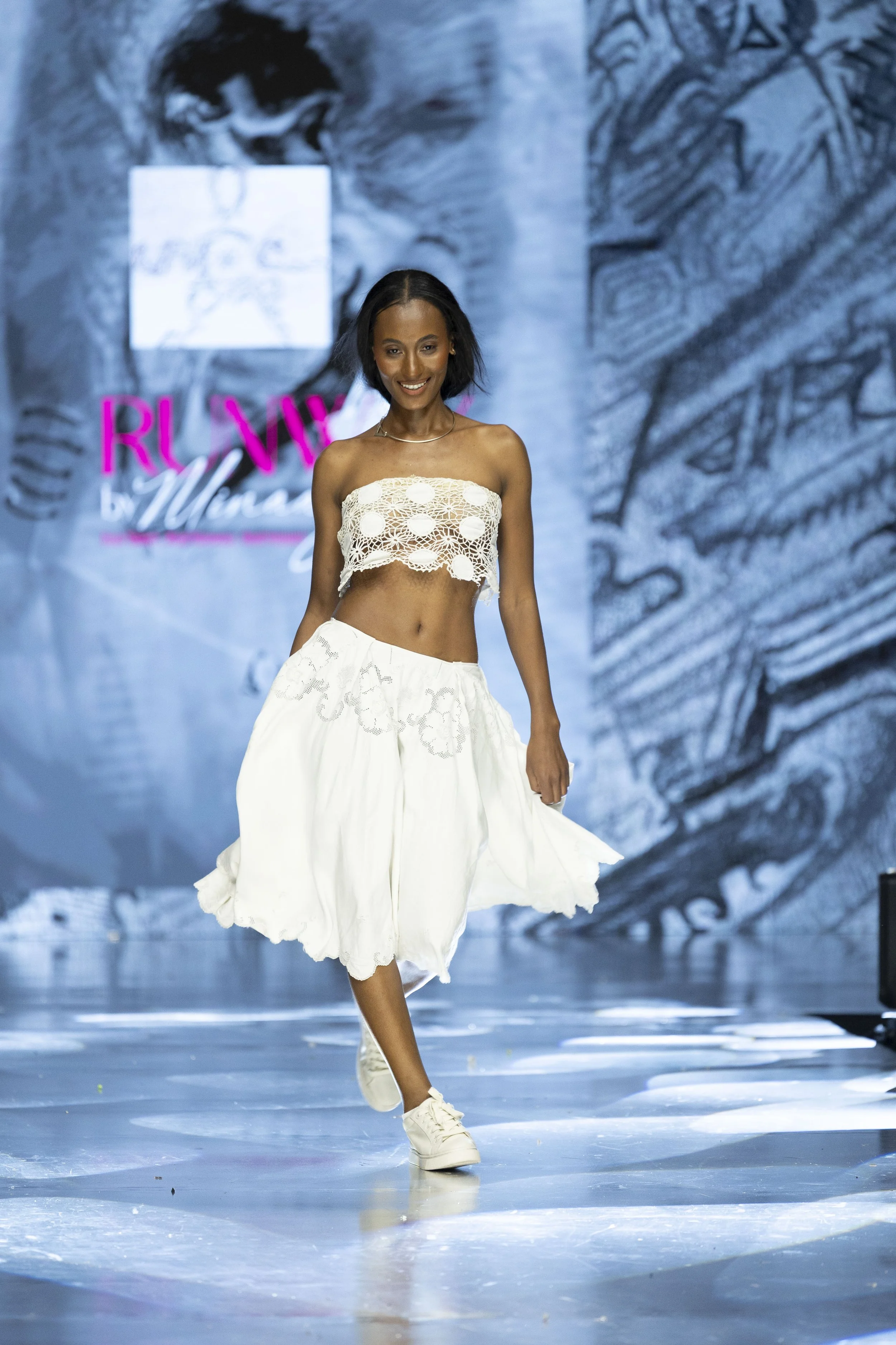 A woman walking on a runway during a fashion show, wearing a white crochet crop top and a flowing white skirt with embroidery, with a large black and white artwork in the background and pink signage.