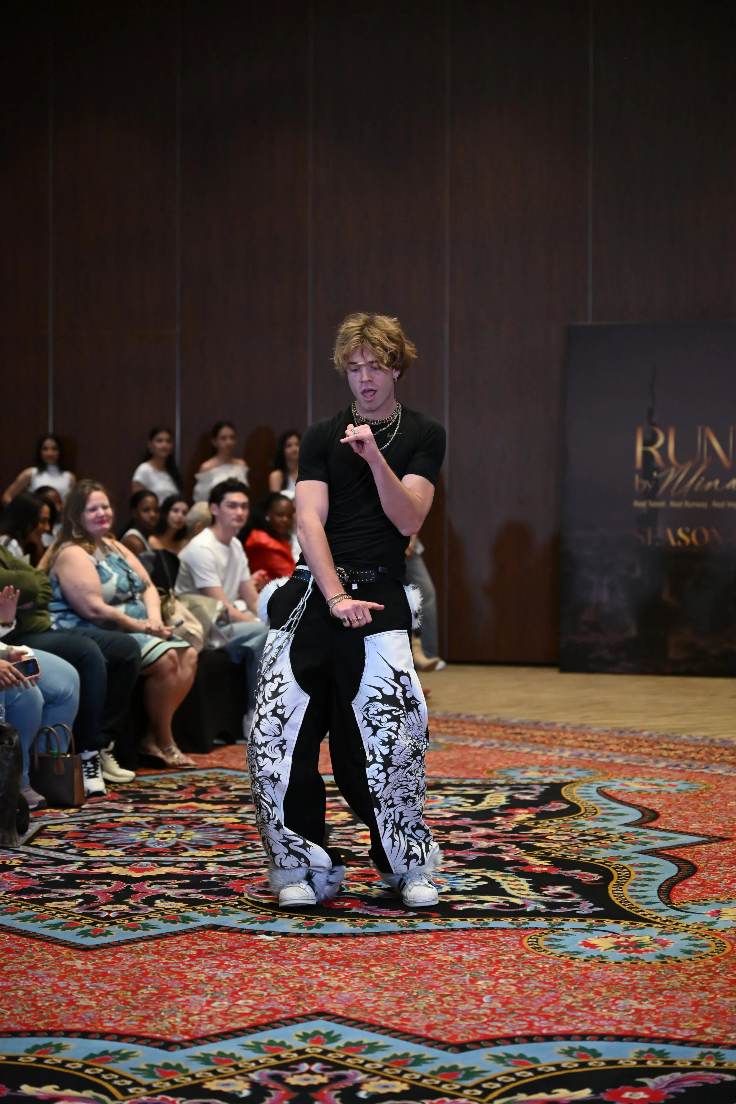 A young man with blond hair is performing or dancing on a patterned carpet at an indoor event, with an audience seated behind him.