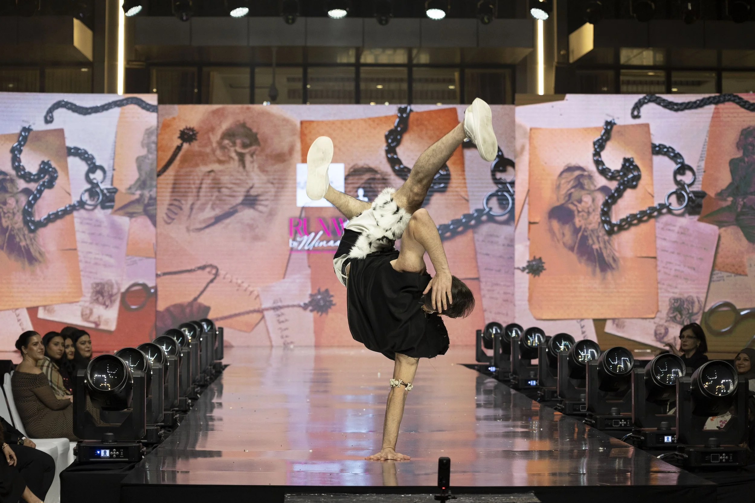 A performer doing a one-handed handstand on a runway, with their legs and arms extended, during a fashion show or event. The background features a large digital screen displaying artistic images and chains, with an audience seated on either side of t