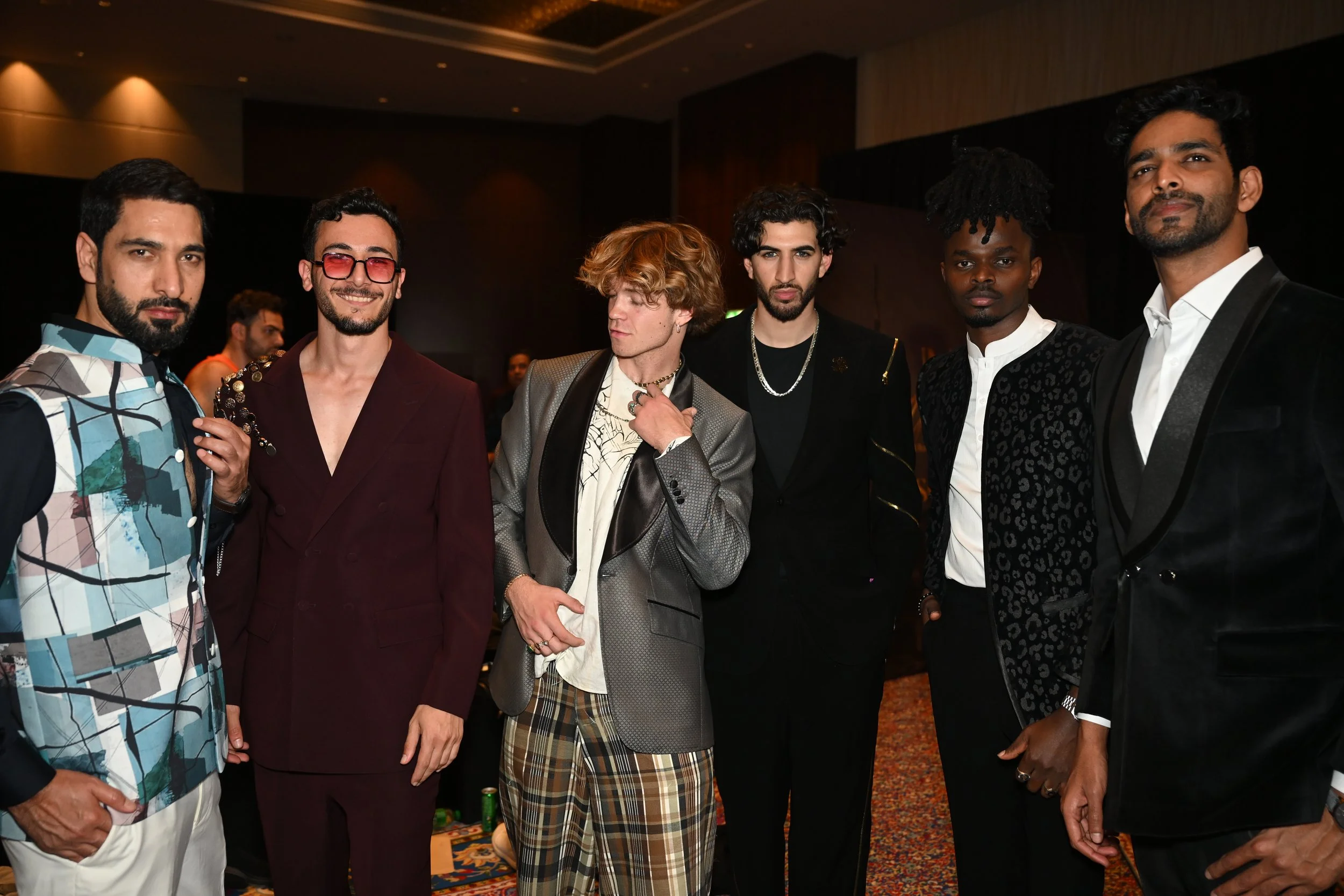 Six men dressed in stylish suits and fashionable clothing standing together indoors at an event.