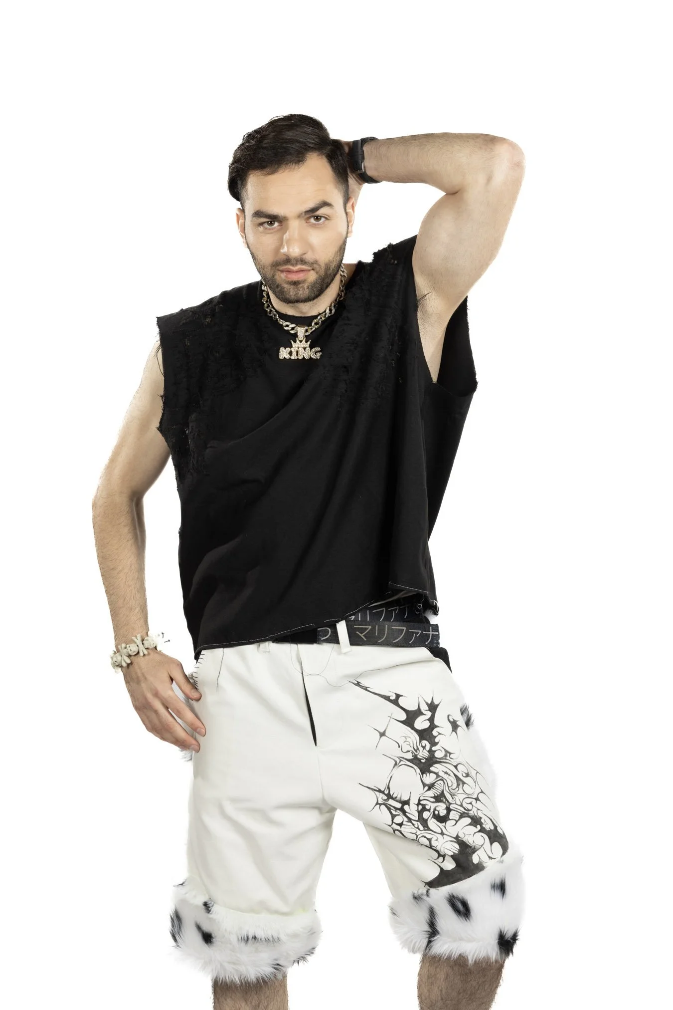 A man with dark hair and a beard posing with one hand behind his head. He is wearing a sleeveless black top with lace details, a gold chain necklace with a "KING" pendant, a bracelet, white shorts with black tribal designs, and fur-lined cuffs at the