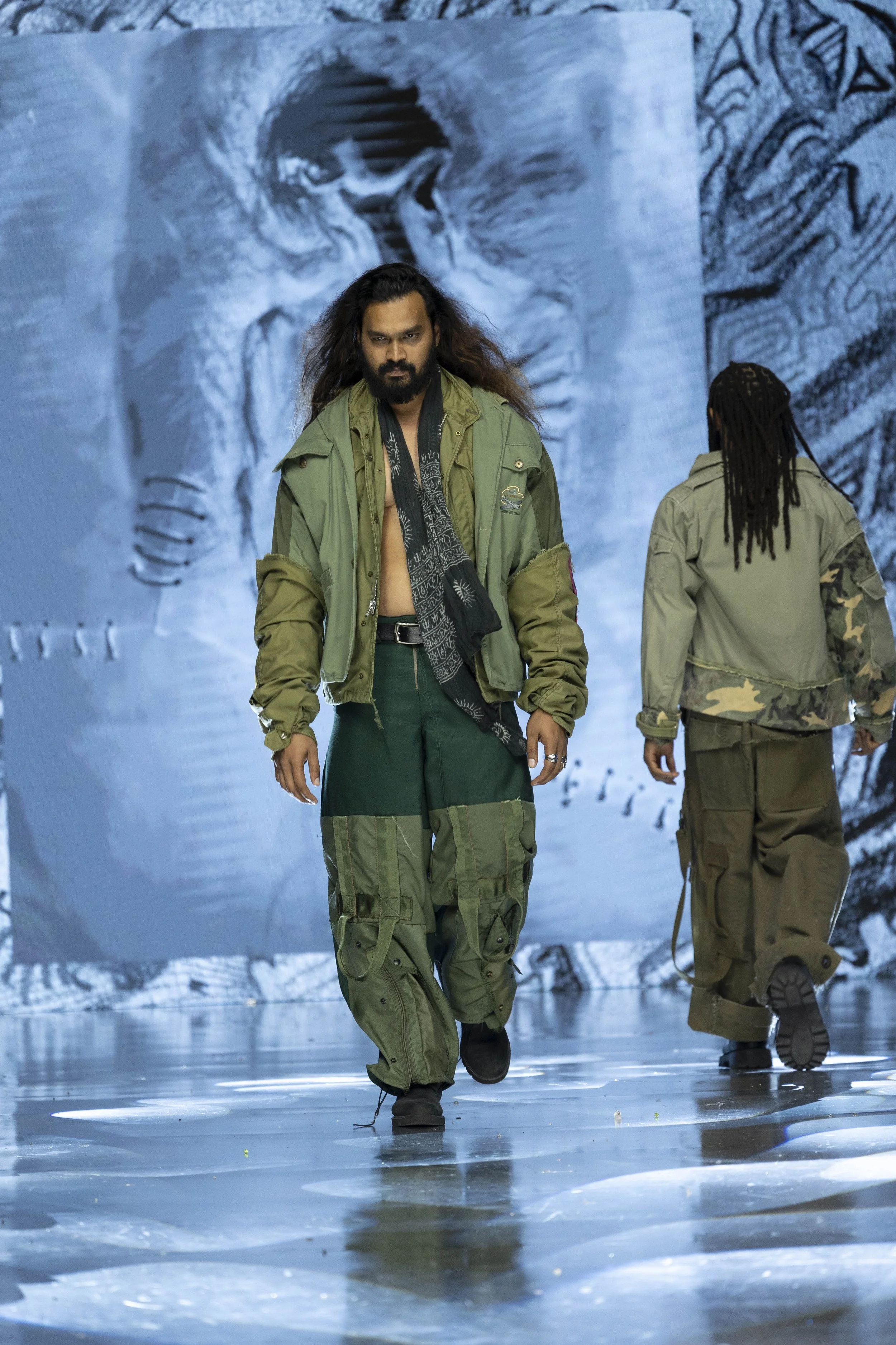 Two models walk on a reflective runway with a large black-and-white artwork of a face in the background, dressed in military-style clothing.