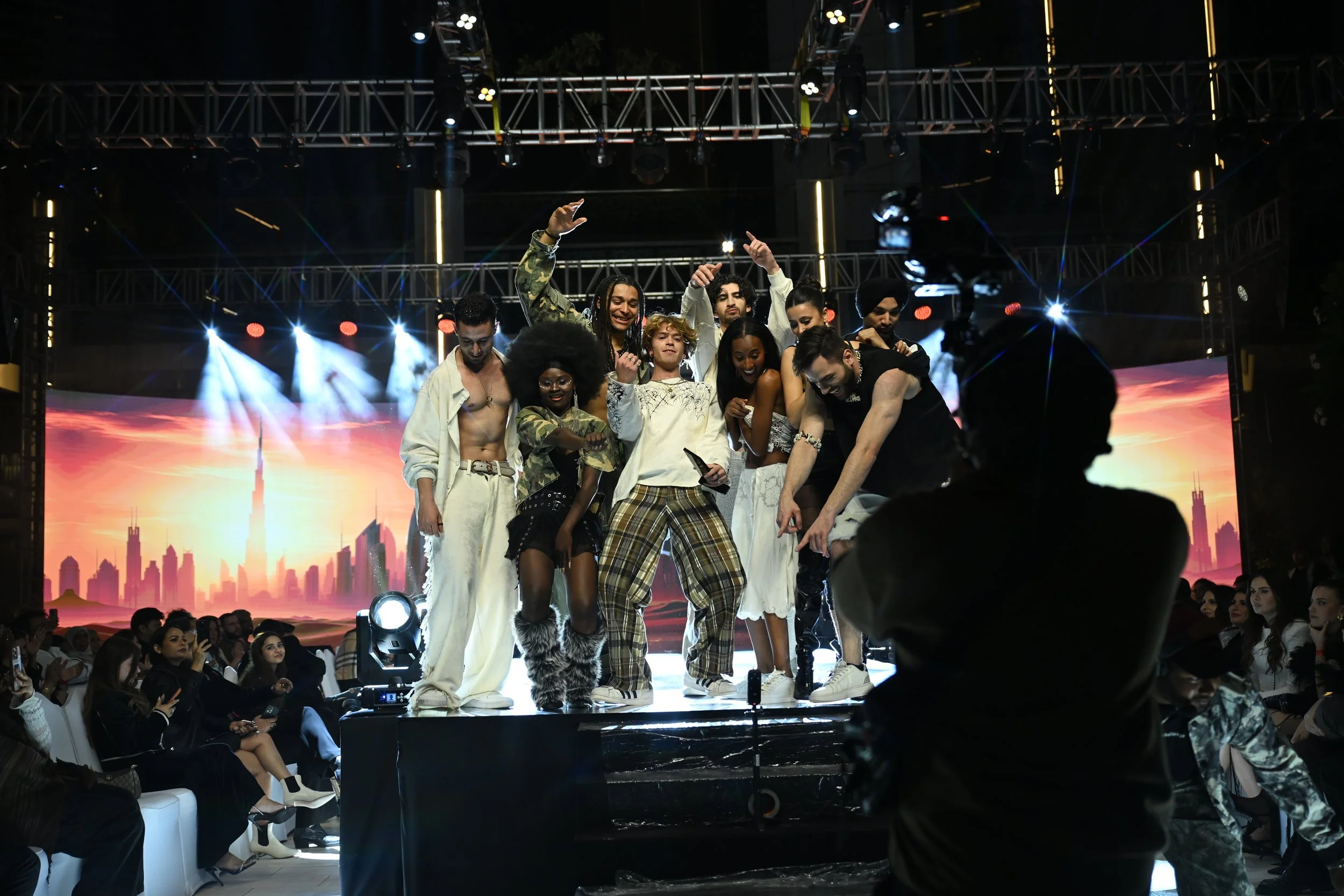 A group of diverse people on a stage at a fashion show or event, with a city skyline and sunset background, and an audience watching.