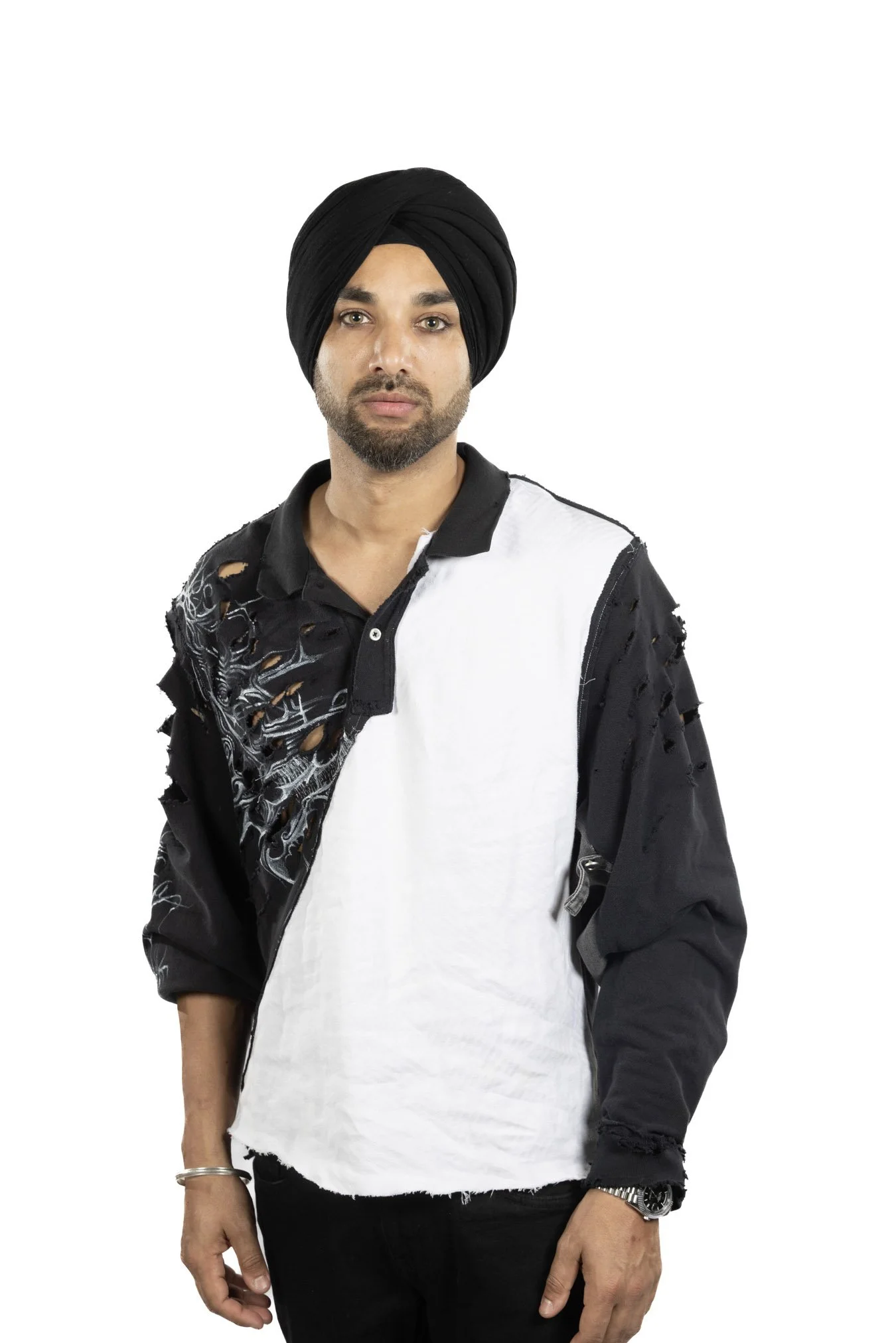 A man with a black turban and beard wearing a black and white distressed jacket over a white shirt, standing against a white background.