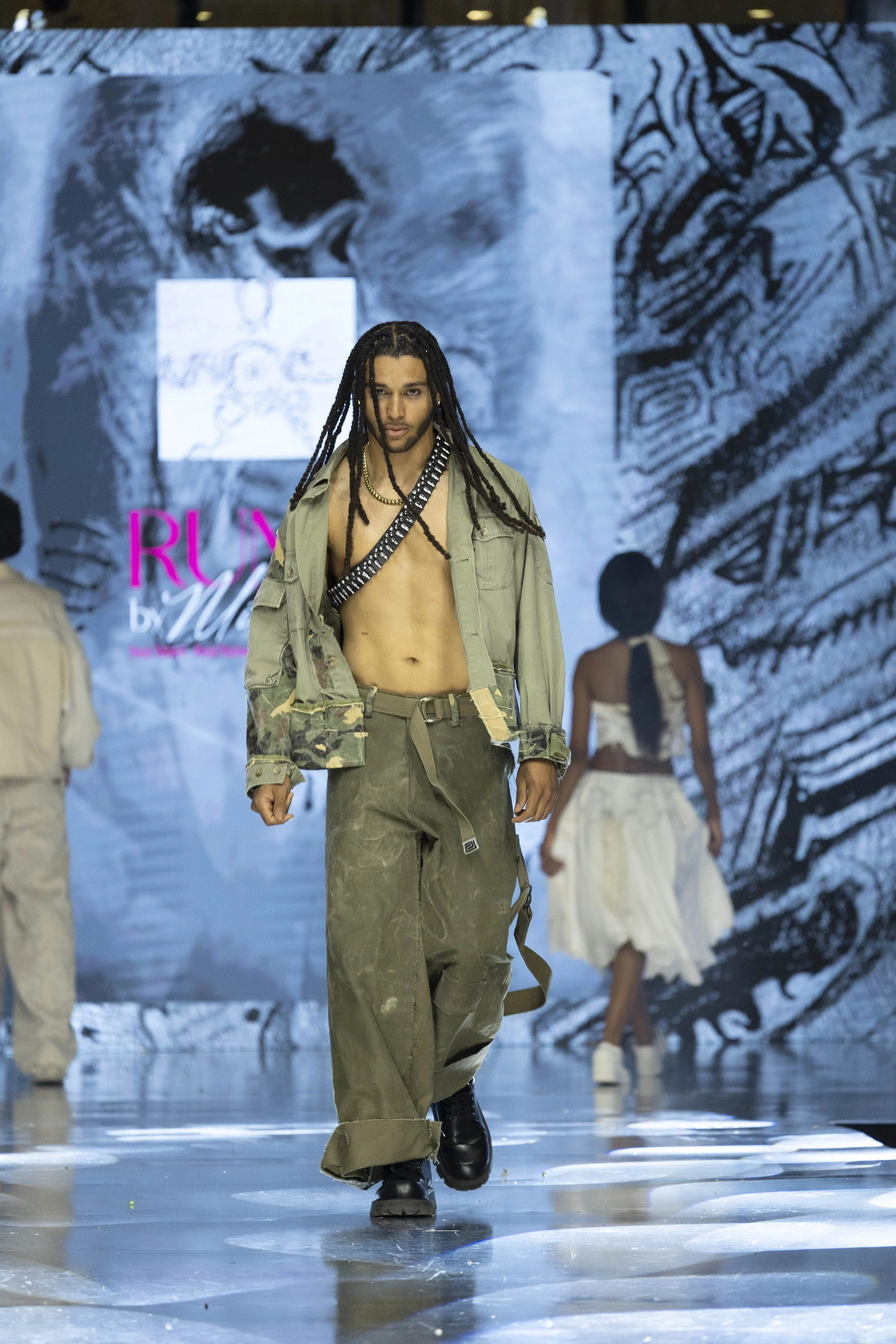 Male model with long dreadlocks walking down a runway in military-style green cargo pants and jacket, shirtless, with a black strap across his chest. Behind him, a woman in a white skirt and top and other models are visible, with a large artistic bla