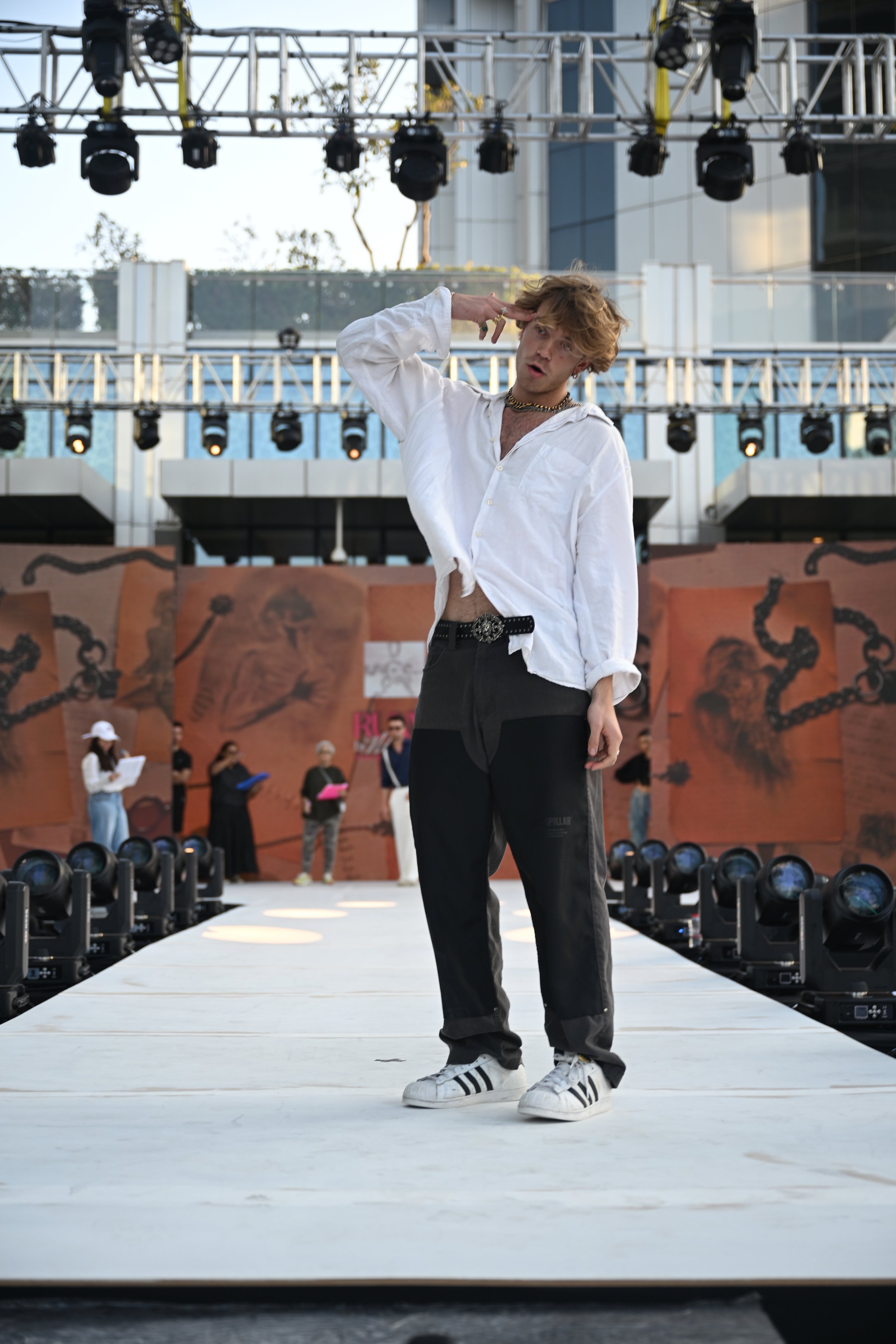 A young man with curly blond hair on a runway stage, wearing a white loose shirt, black jeans, and white Adidas sneakers. He is saluting the audience with his right hand, standing in front of a background with painted images of chains and a brain. Se