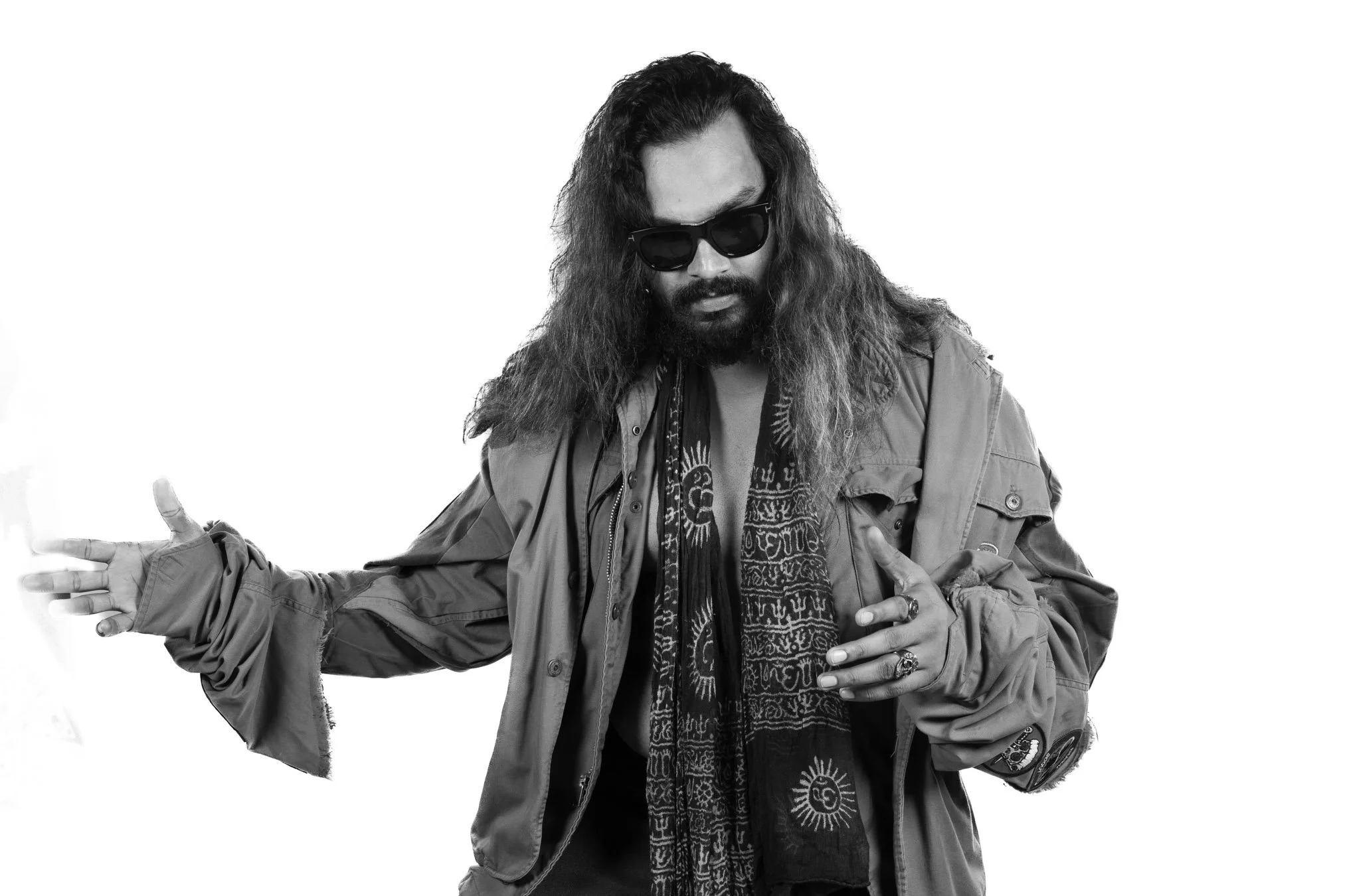 Black and white photo of a man with long curly hair, wearing sunglasses, a patterned scarf, and a layered jacket, posing with his arms outstretched.